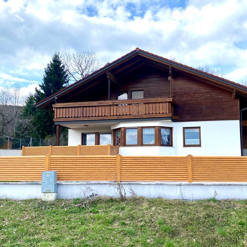 Ein zweistöckiges Haus mit brauner Holzfassade und Balkon, umgeben von einem Holzzaun, in einem grün bewachsenen Bereich mit Bäumen im Hintergrund.