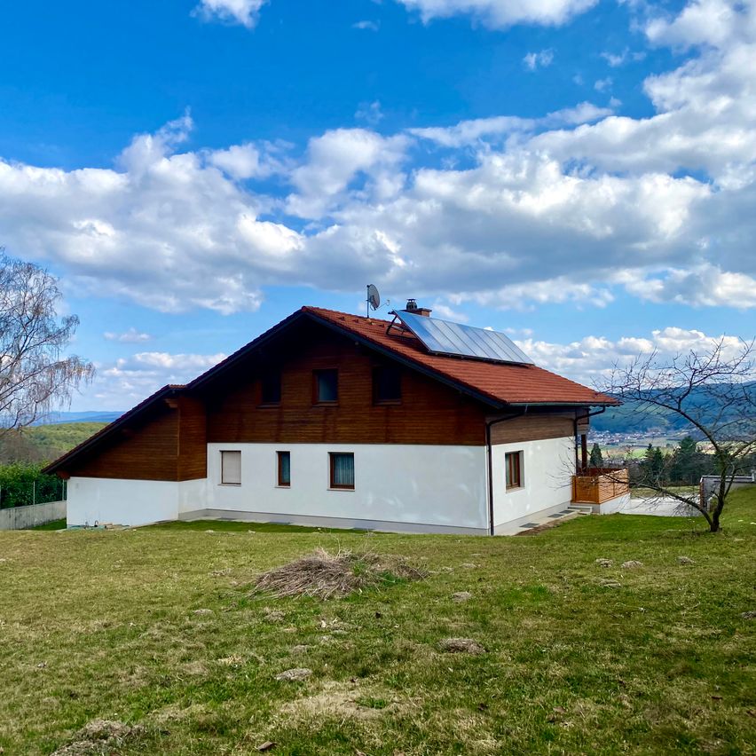 Ein modernes Haus mit Solarmodulen auf einem Hügel, umgeben von üppiger Grünfläche und einem klaren blauen Himmel mit verstreuten Wolken.