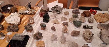 Ein mit einem weißen Tuch bedeckter Tisch zeigt verschiedene Steine und Mineralien. Einige stehen auf Ständern mit Etiketten. Hinter dem Tisch befinden sich zwei Holzstühle und eine Wand mit einer Zeichnung.