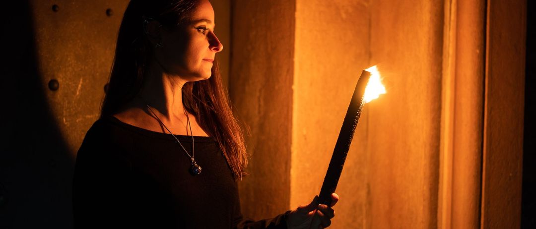 Eine Frau in einem dunklen Raum hält eine brennende Fackel, trägt ein schwarzes Kleid und eine Halskette. Das Licht der Fackel beleuchtet ihr Gesicht und die Wand hinter ihr.