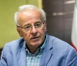 Ein älterer Mann mit weißen Haaren und Brille sitzt in einem Büro und trägt einen blauen Anzug über einem karierten Hemd. Er scheint zu sprechen oder ein Interview zu geben.