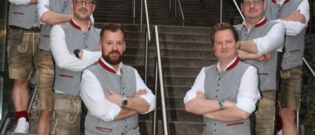 Sieben Männer in traditioneller bayerischer Kleidung posieren auf den Stufen eines Gebäudes. Sie haben alle die Arme verschränkt.