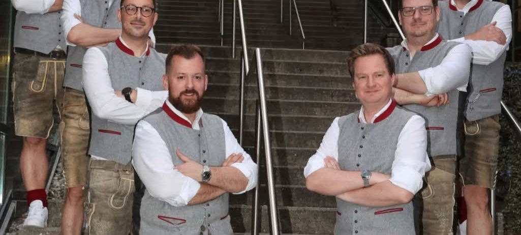 Sieben Männer in traditioneller bayerischer Kleidung posieren auf den Stufen eines Gebäudes. Sie haben alle die Arme verschränkt.