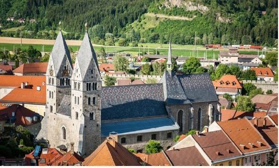 Luftaufnahme einer historischen Stadt mit einer großen Kirche und Terrakotta-Dächern, eingebettet zwischen grünen Hügeln und einem üppigen grünen Tal.