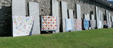 Eine Reihe bunter Quilts hängt vor einer Steinmauer an einer Leine, mit Fahnen und einem Metallgeländer darüber.