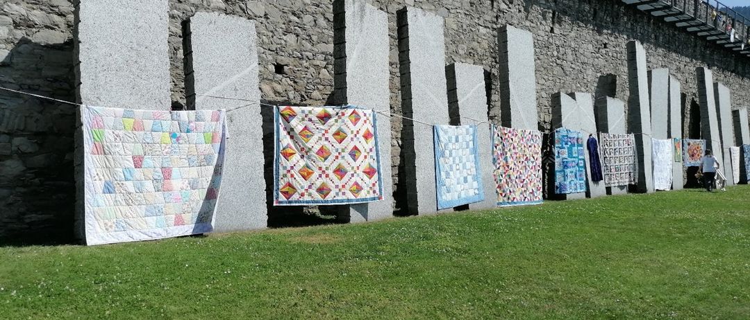 Eine Reihe bunter Quilts hängt vor einer Steinmauer an einer Leine, mit Fahnen und einem Metallgeländer darüber.
