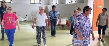 Eine Gruppe von Frauen geht in einem Raum mit gelbem Boden, möglicherweise beim Training. Im Raum stehen Stühle und Tische.