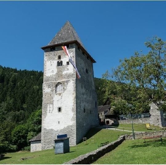 Ein mittelalterlicher Turm mit einer Flagge auf der Spitze steht auf einer Rasenfläche mit einem Baum. Eine Bank und ein Pfahl befinden sich auf der rechten Seite. Der Turm ist von Bäumen und Bergen umgeben.