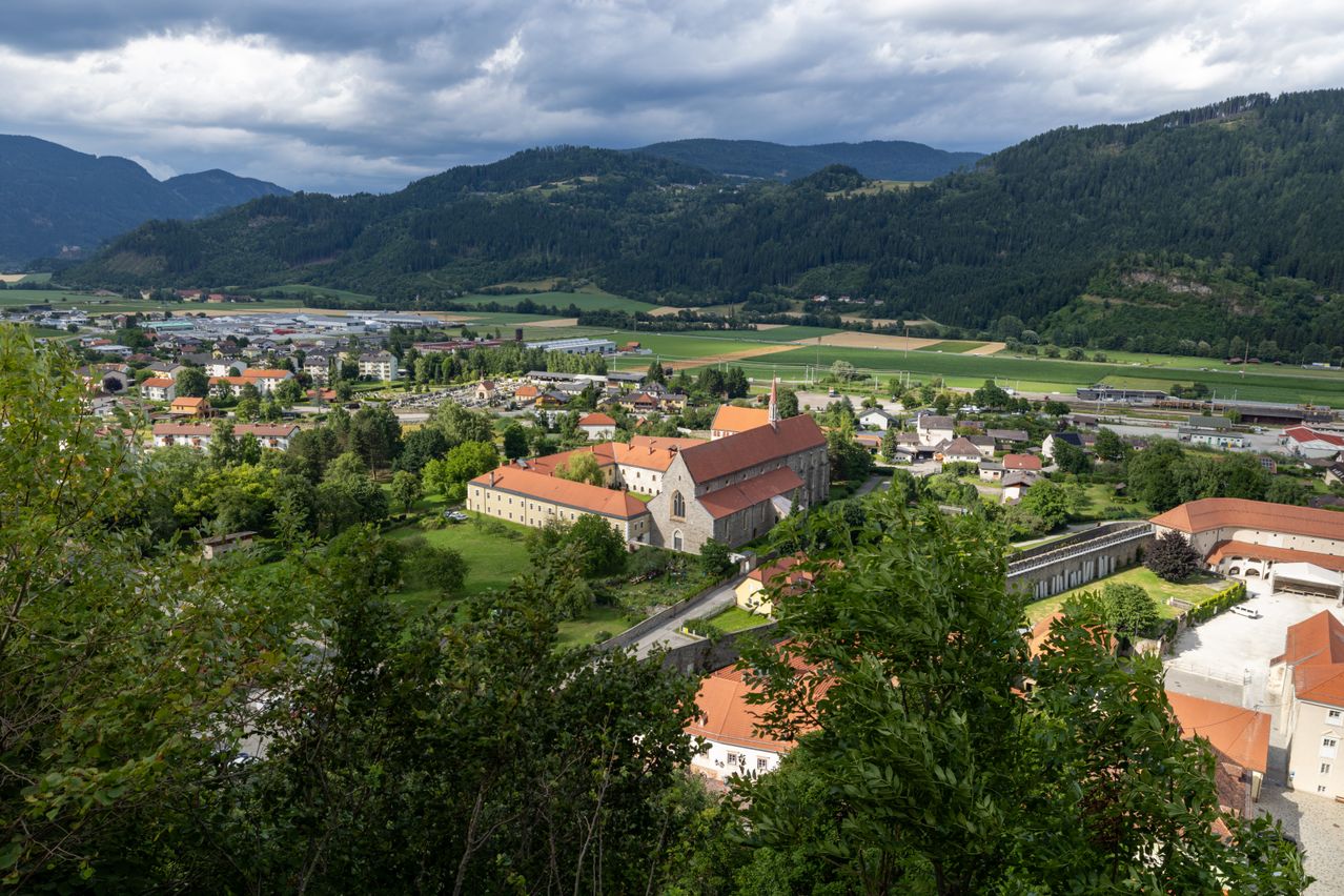 Eine Luftaufnahme einer Stadt mit einer Kirche und Bergen. Gebäude, Bäume und ein grünes Feld sind unter einem bewölkten Himmel sichtbar.