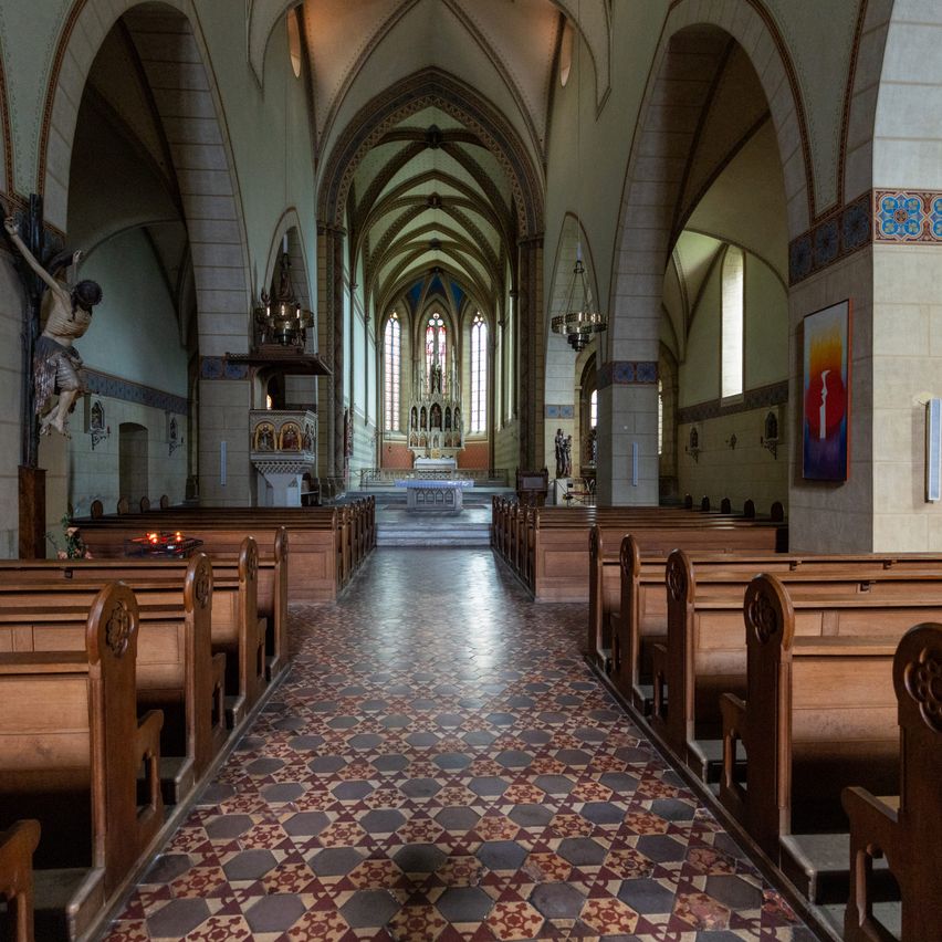 Eine leere Kirche mit hölzernen Kirchenbänken, einem Altar und einem Kreuz an der Wand. Ein farbenfroher Boden und dekorative Elemente schmücken die Wände.