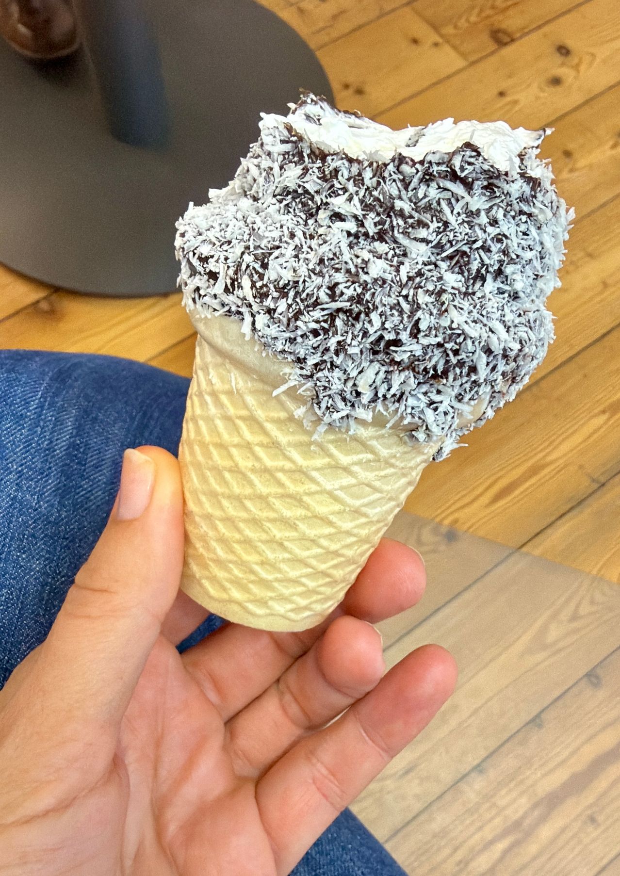 A hand holds an ice cream cone with a topping of white coconut shavings and chocolate sprinkles on a wooden floor.