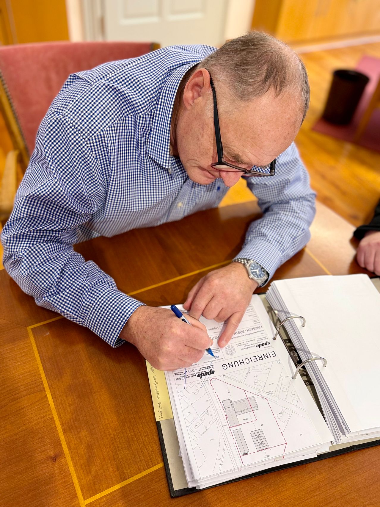 A man wearing glasses is sitting on a chair and writing on a document with a pen. A folder with the word 'ENGINEERS' is open on the desk.