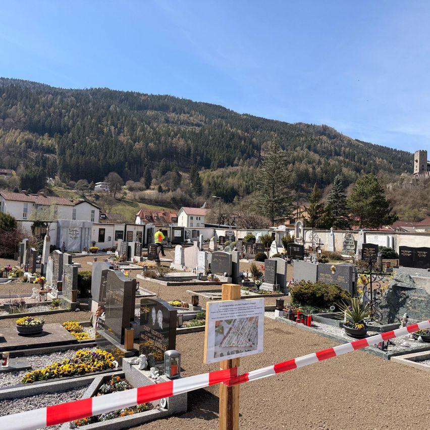 A cemetery is surrounded by a barrier tape with a sign. It has many tombstones and flowers. A worker is cleaning the area. In the distance, there are buildings and a mountain.