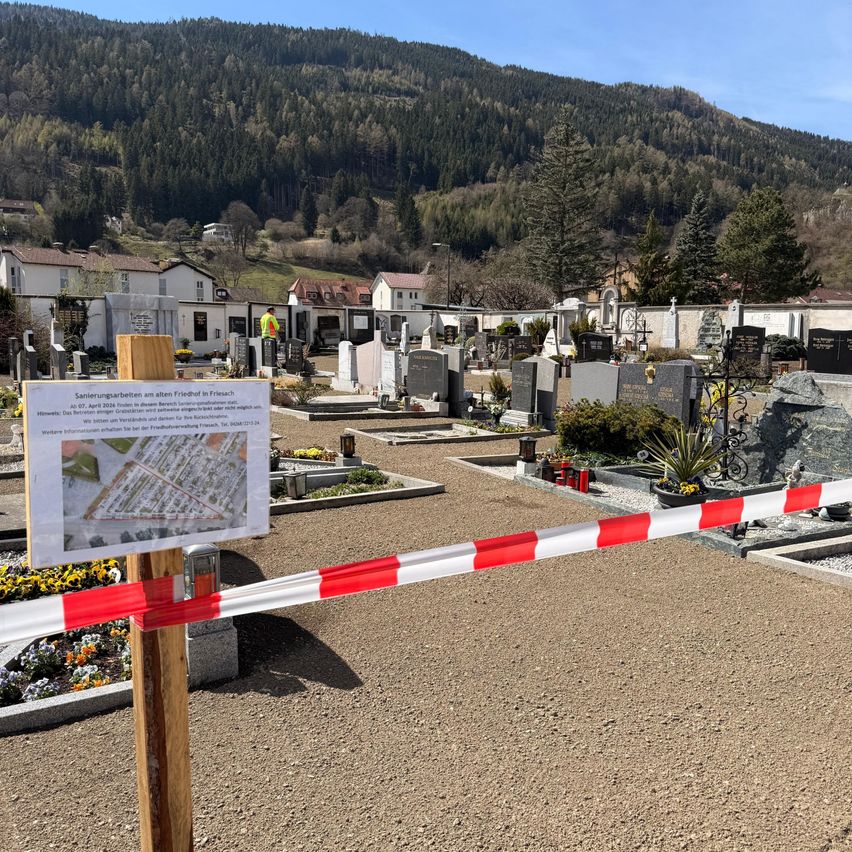 A cemetery with several tombstones and a map attached to a wooden post. There is a red and white ribbon barrier in front. In the background are houses and a mountain.