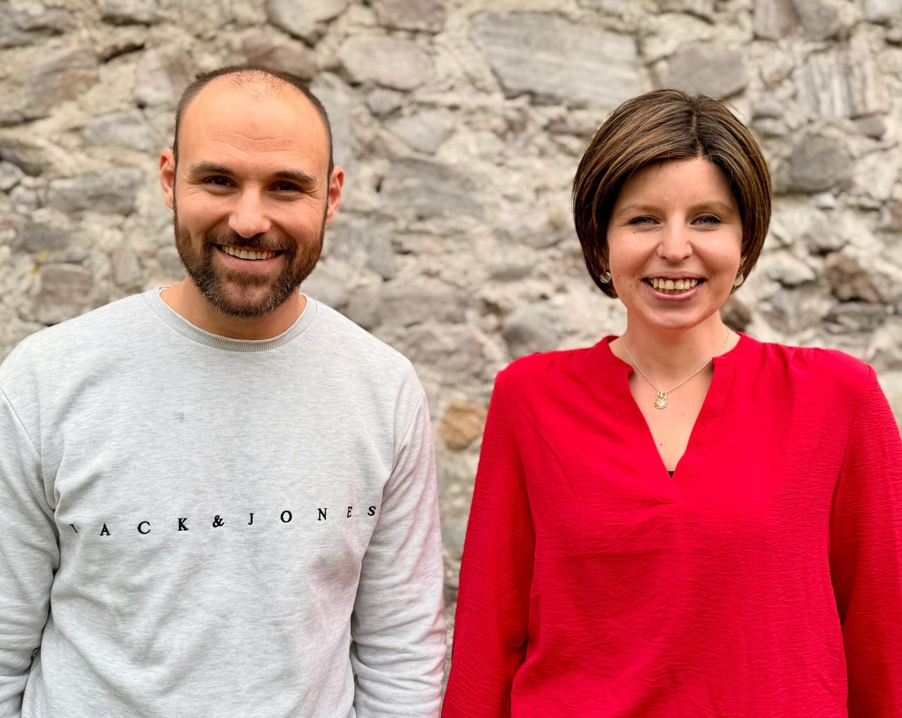A man and a woman smiling at the camera. The man is wearing a gray shirt with black letters, and the woman is wearing a red blouse. They are standing against a stone wall.