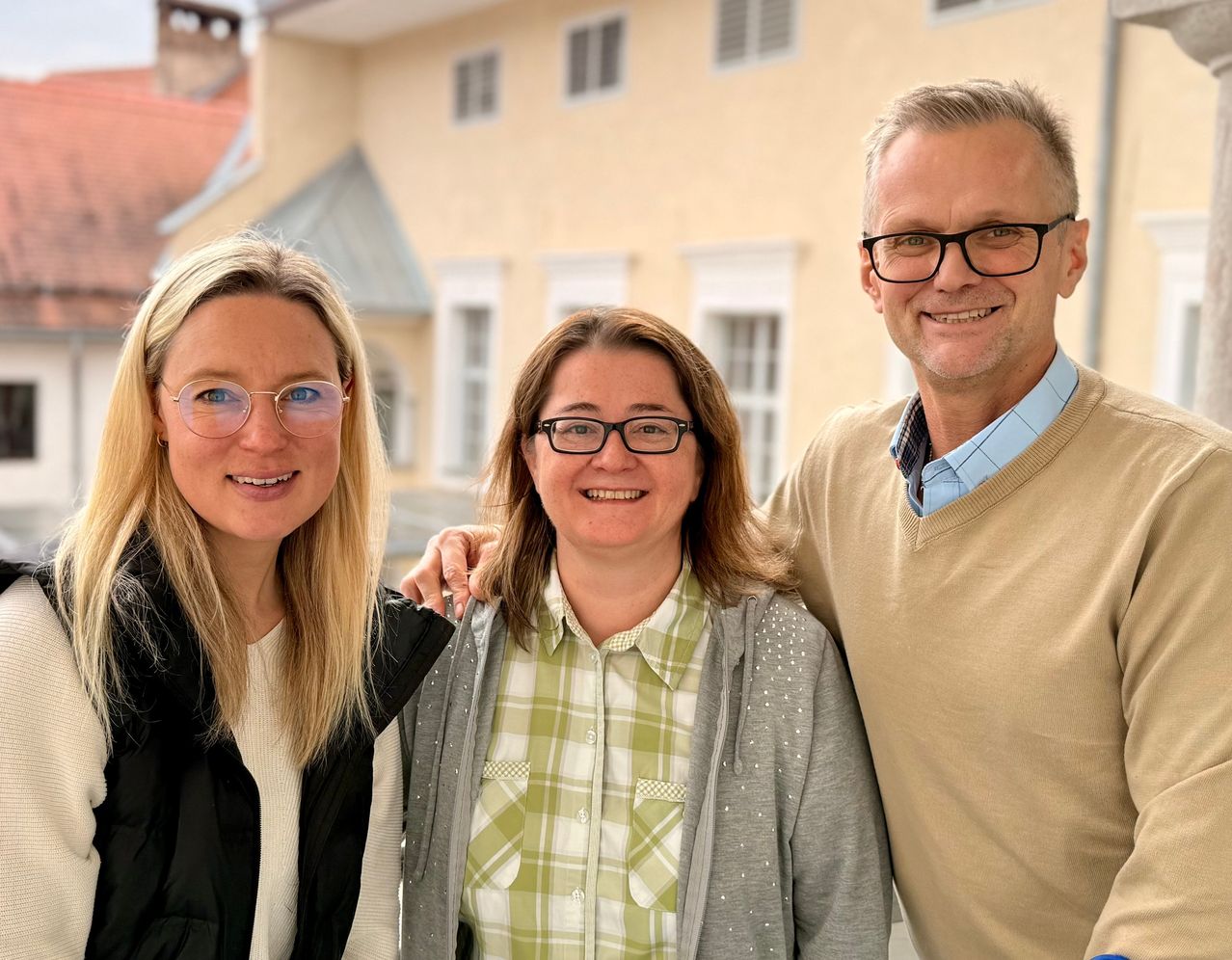 Drei Menschen stehen vor einem Gebäude. Zwei Frauen und ein Mann, alle lächeln. Die Frau links hat lange blonde Haare und eine Brille. Die Frau in der Mitte hat kurze braune Haare und eine Brille. Der Mann rechts hat eine Brille und einen beigen Pullover.