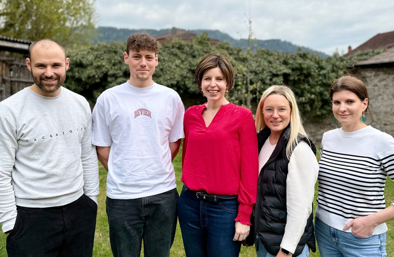 Eine Gruppe von fünf Personen steht in einem Grasfeld mit einem Hintergrund aus grünen Büschen und Bergen. Ein Mann in einem weißen T-Shirt lächelt, während eine Frau in einem roten Oberteil und eine andere Frau in einer schwarzen Weste ebenfalls lächeln.