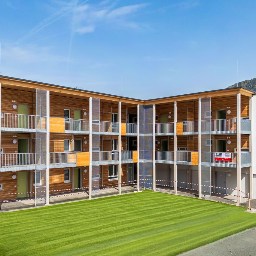 A modern apartment building with multiple floors, wooden exteriors, and balconies. The building is surrounded by a large green lawn under a clear blue sky.