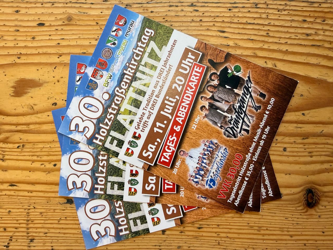 Ein Stapel von Broschüren, die ein Ereignis mit dem Datum 12. Juli 2020 auf einem Holztisch bewerben. Der Text auf der Broschüre ist auf Deutsch.