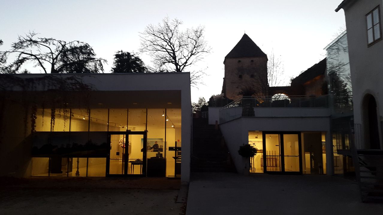 Das Gebäude hat Glastüren und Fenster mit eingeschalteten Lichtern. Eine Treppe führt zu einem Turm. Bäume sind im Hintergrund.