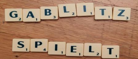 Scrabble-Steine, die 'GABITZ' und 'SPIELT' auf einer Holzoberfläche bilden. Jedem Stein ist eine Nummer zugeordnet.