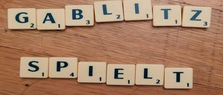 Scrabble-Steine bilden die Wörter 'GABLIZ' und 'SPIELT' auf einem Holztisch. Zahlen zeigen den Wert jedes Buchstabens an.