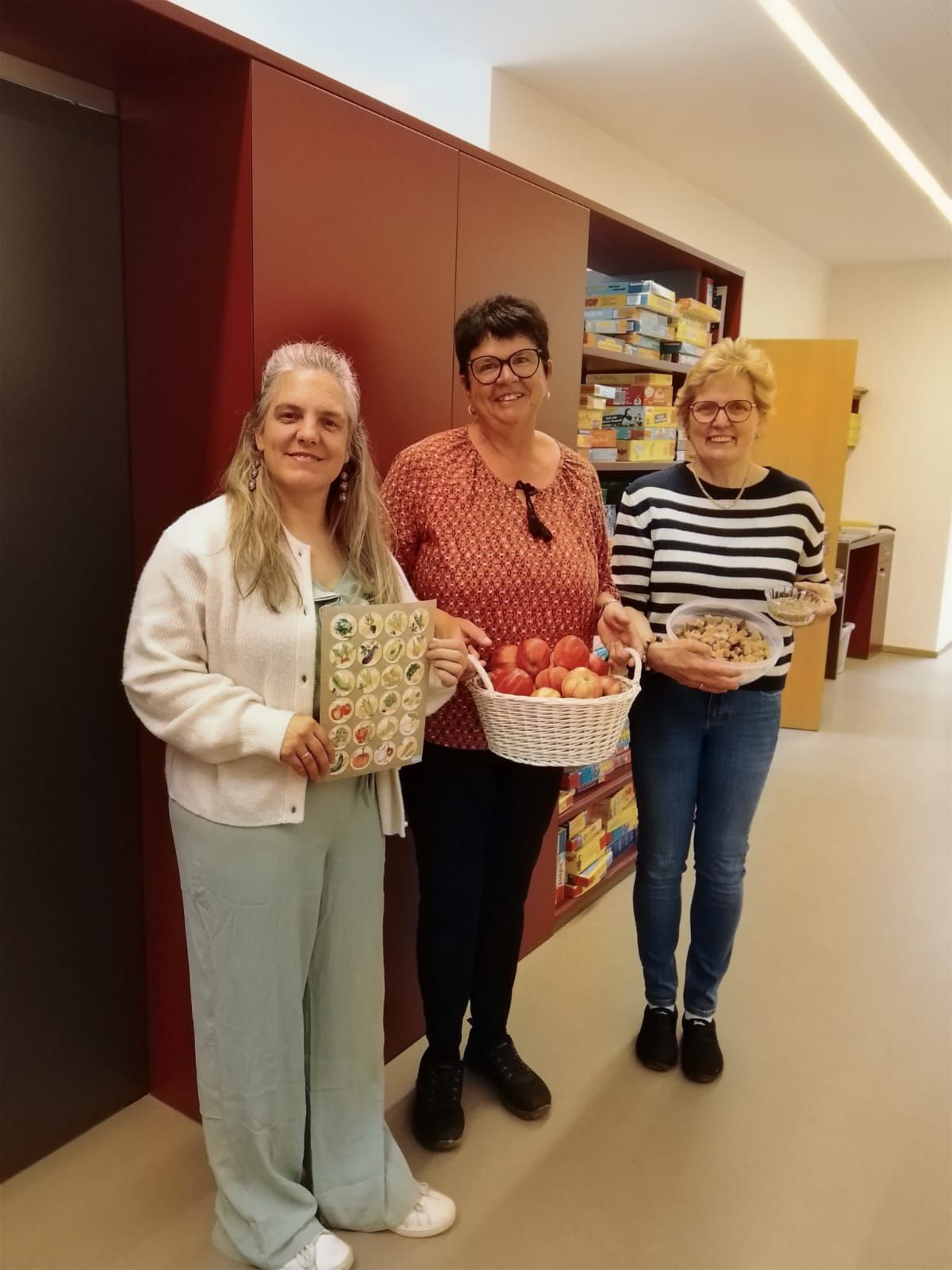 Three women standing in a room, smiling and holding baskets. One has a box of fruits, another has a basket of tomatoes, and the last has a bowl of food.
