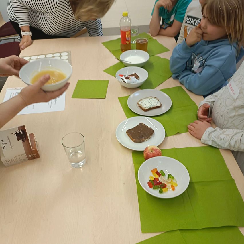 A group of people are seated around a table, preparing food. One person holds a bowl with yellow food. Various plates of food and drinks are set up on the table.