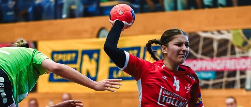 Zwei Frauen spielen Handball; eine springt, um den Ball zu fangen. Das Spielfeld ist mit Linien markiert, und Zuschauer beobachten von den Seiten.