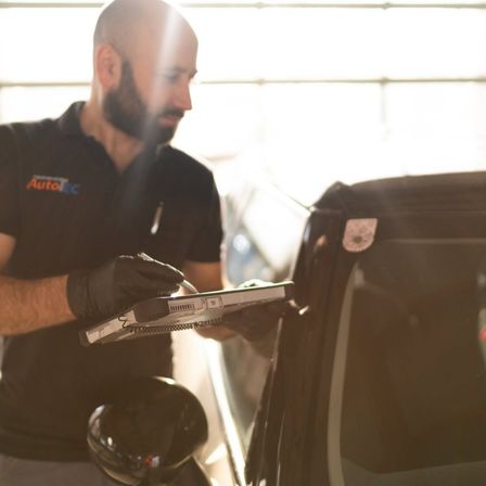 Ein Mann in einem schwarzen Shirt mit 'AutoEC', der Handschuhe trägt, arbeitet mit einem Laptop an einem Auto.