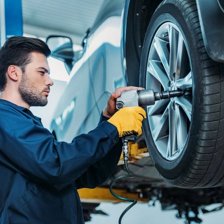 Ein Mann in einer blauen Uniform und gelben Handschuhen benutzt ein Werkzeug, um die Radmuttern eines Autos festzuziehen.