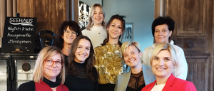 Eine Gruppe von Frauen steht in einem Raum, lächelt und posiert für ein Foto. Sie tragen verschiedene Outfits. Ein Schiefertafel mit den Worten 'SEEHAUS' ist an der Wand. Ein Kühlschrank steht links, ein Schrank rechts. Die Holztür ist hinter ihnen.