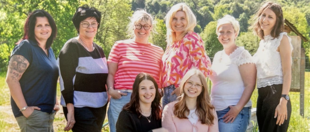 Eine Gruppe lächelnder Frauen unterschiedlichen Alters steht und sitzt auf einer Bank in einem Grasfeld mit malerischer Kulisse. Sie sind in verschiedenen lässigen Outfits gekleidet.