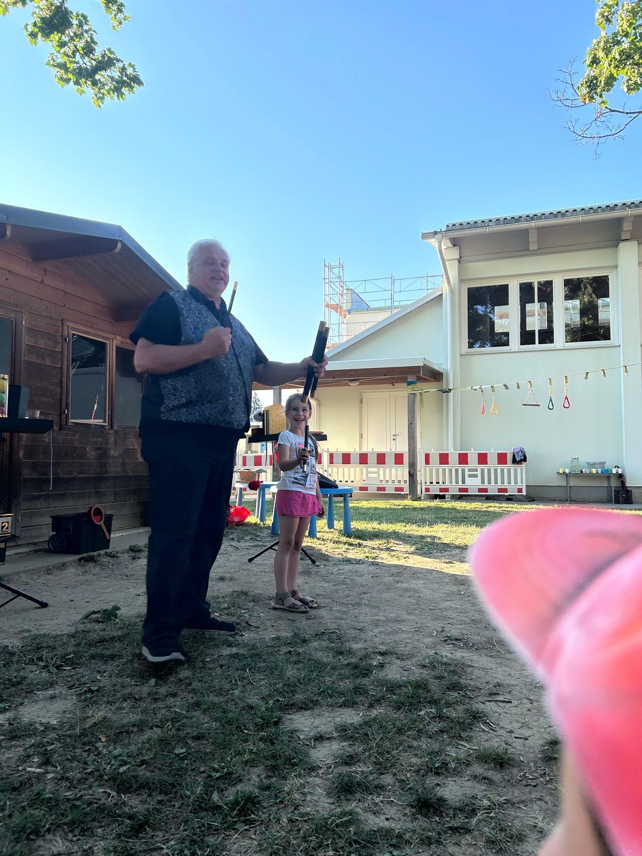 A man and a young girl stand in a yard, the man holding a stick and smiling, the girl holding a stick and looking at him. Behind them, there are two houses, one with a wooden exterior and the other with a white exterior.