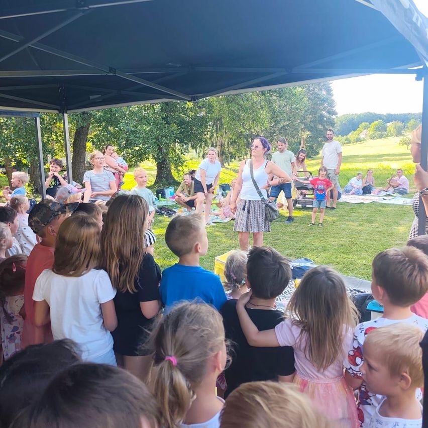 A group of children and adults gather under a tent in a park, with a woman speaking to them. Some people sit on the grass while others stand around. Trees and a distant field are visible.