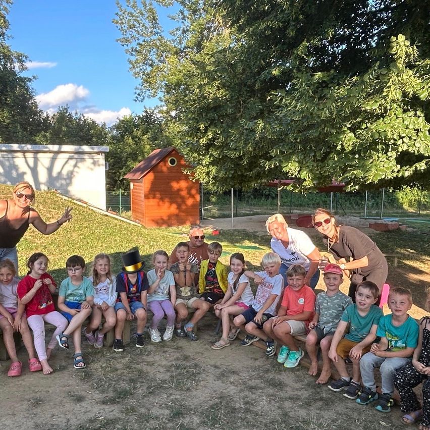 A group of children and adults are sitting on a sloped area, possibly in a park. They are wearing casual clothing. Behind them are trees, a small wooden house, and a white structure.