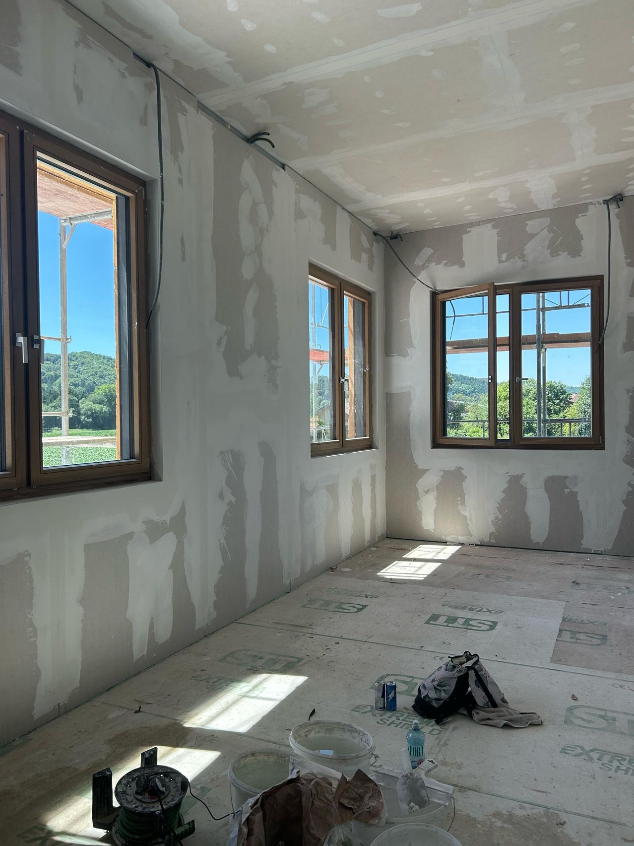 Ein leerer Raum im Bau mit unfertigen Wänden, drei Fenstern und Sonnenlicht. Einige Gegenstände liegen auf dem Boden.