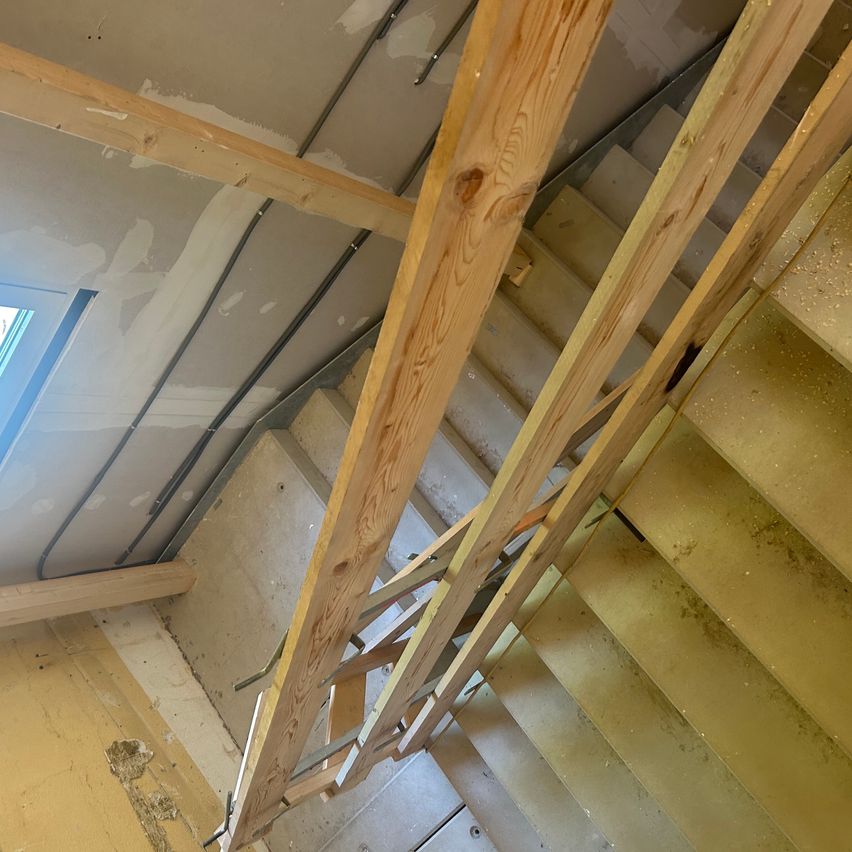 Ein Innenraum eines Hauses zeigt die Holzstruktur der Treppe mit einigen unfertigen Teilen. Die Stufen sind aus Holz und haben eine gelbe Umrandung.