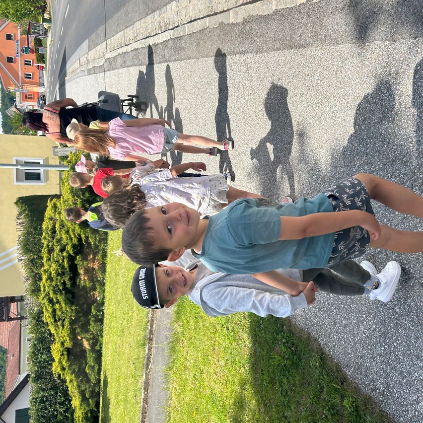 Eine Gruppe von Kindern und Erwachsenen läuft auf einer Straße mit Kies, einige tragen Hüte, neben einem Rasenbereich und Gebäuden im Hintergrund.