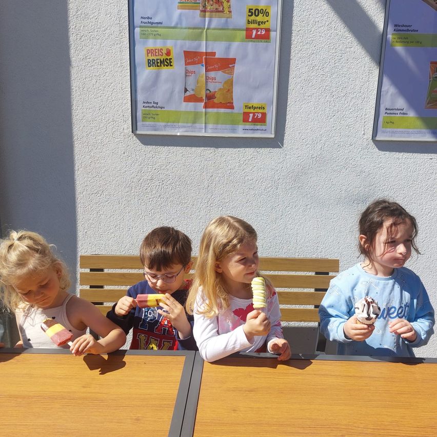 Vier Kinder sitzen mit Eis am Stiel am Tisch. Ein Plakat im Hintergrund bewirbt Kartoffelchips und andere Snacks.