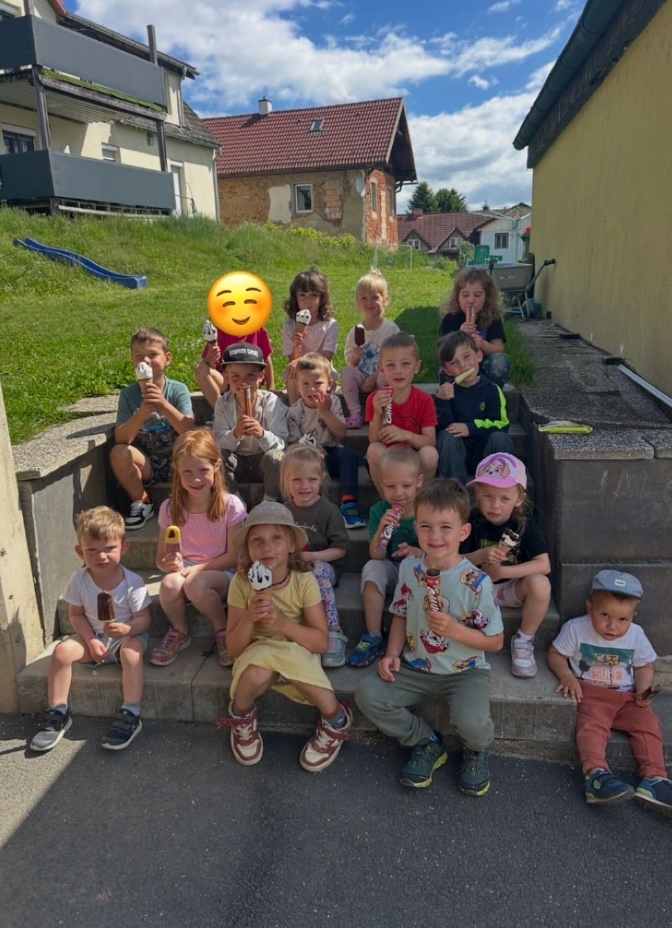 Eine Gruppe von Kindern sitzt auf Stufen und hält Eis am Stiel, lächelt für ein Foto. Ein gelbes Smiley-Gesicht ist in der Mitte, mit Häusern und einem Grashügel im Hintergrund.