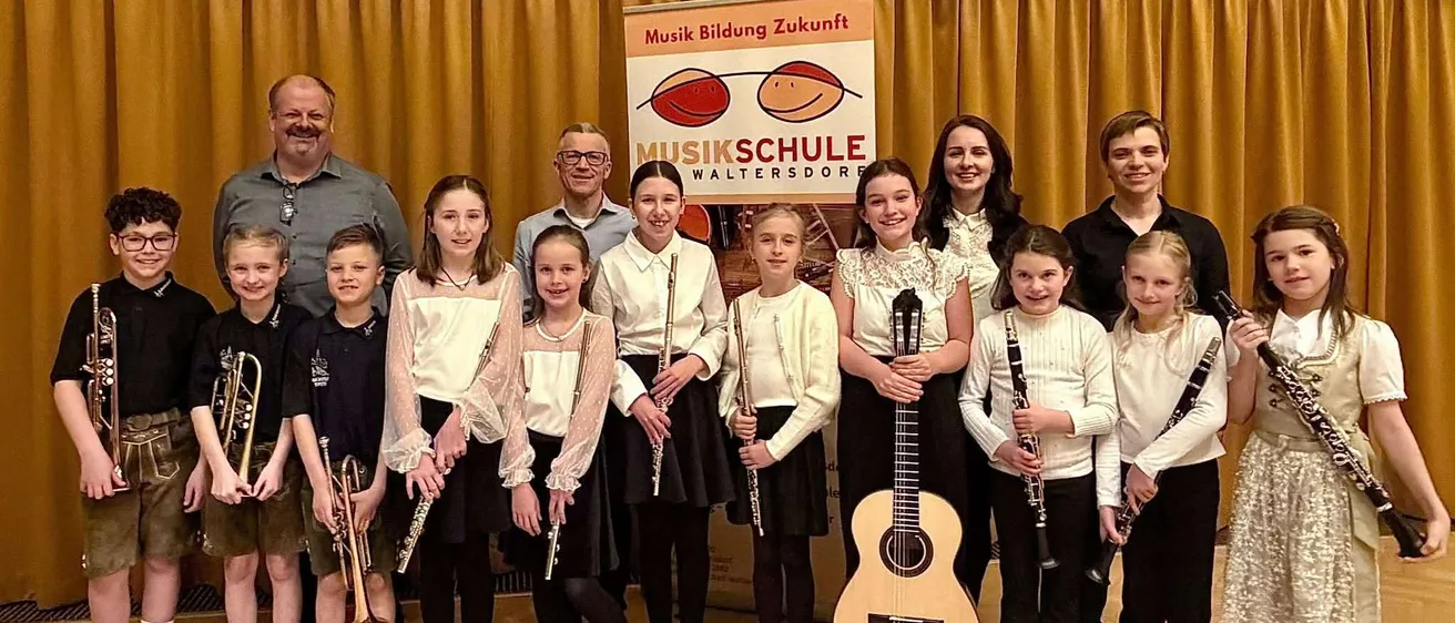 Eine Gruppe von Kindern und Erwachsenen steht zusammen mit Musikinstrumenten vor einem Schild, das Musikschule Waltersdorre besagt.