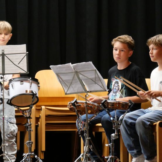 Drei junge Jungen sitzen und spielen Trommeln, jeder hält Trommelstöcke. Sie stehen vor Notenständern mit Notenblättern. Der Junge auf der linken Seite schaut auf seine Musik, während die anderen beiden sich auf ihre Trommeln konzentrieren.