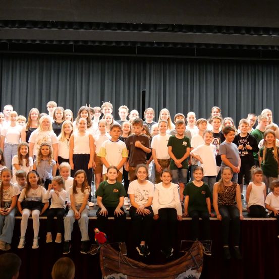 Eine Gruppe von Kindern und Jugendlichen in verschiedenen Farben, die auf einer Bühne mit schwarzem Vorhang sitzen und stehen.