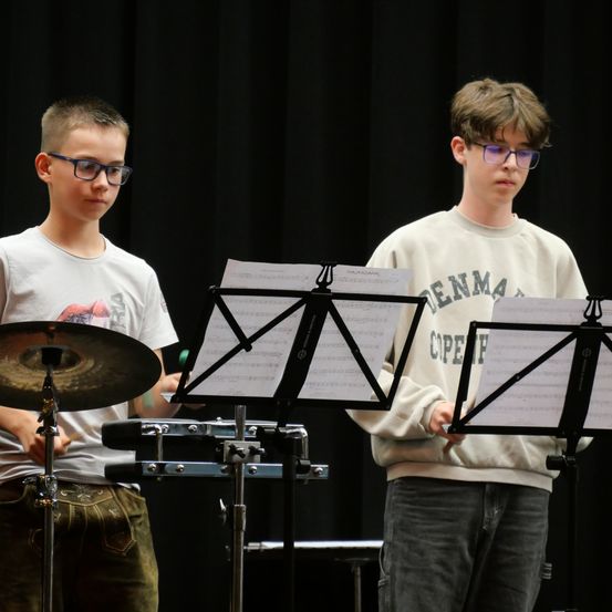 Zwei junge Jungen stehen vor Notenständern, einer hält einen Drumstick und der andere ein Schlagzeug. Beide tragen eine Brille und scheinen sich in einem Musikraum zu befinden.