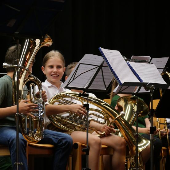 Eine Gruppe von Kindern sitzt auf Stühlen und spielt Blechblasinstrumente. Ein Mädchen vorne lächelt und hält eine Tuba. Notenblätter stehen vor ihnen auf Ständern.