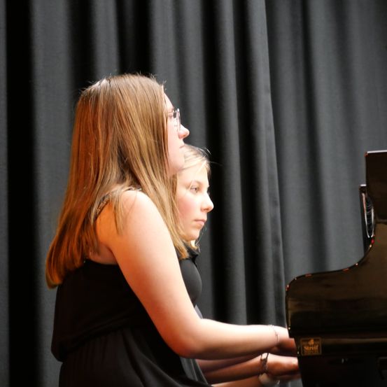 Zwei junge Mädchen in schwarzen Kleidern spielen Klavier auf der Bühne. Das Mädchen vorne trägt eine Brille und schaut auf die Klaviertasten.