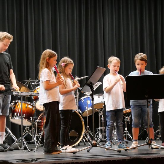 Fünf Kinder stehen auf der Bühne, jedes hält eine Blockflöte. Sie sind in Freizeitkleidung gekleidet und scheinen bei einem Schulkonzert aufzutreten. Hinter ihnen sind verschiedene Musikinstrumente aufgebaut, einschließlich Trommeln und Becken.