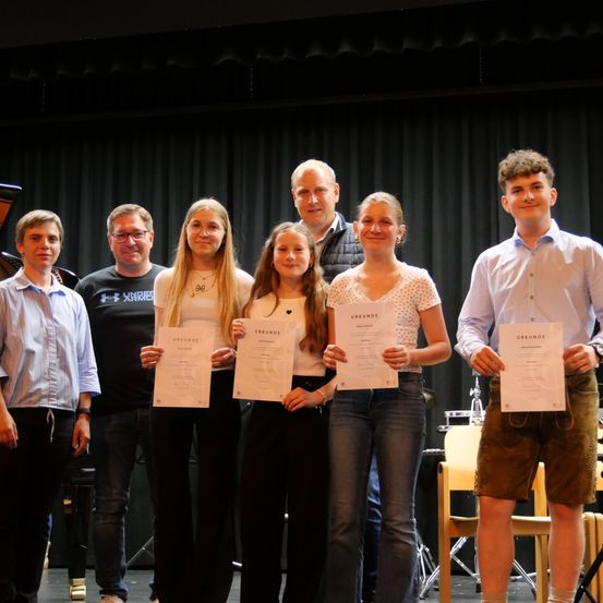 Eine Gruppe junger Menschen und Erwachsene stehen auf einer Bühne und halten Urkunden. Sie lächeln und posieren für ein Foto. Die Bühne hat einen schwarzen Hintergrund und ein Klavier auf der linken Seite.