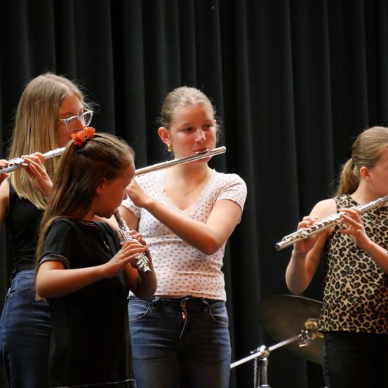 Vier junge Mädchen spielen Flöten auf der Bühne, wobei ein Mädchen in der Mitte eine Blume in den Haaren trägt.